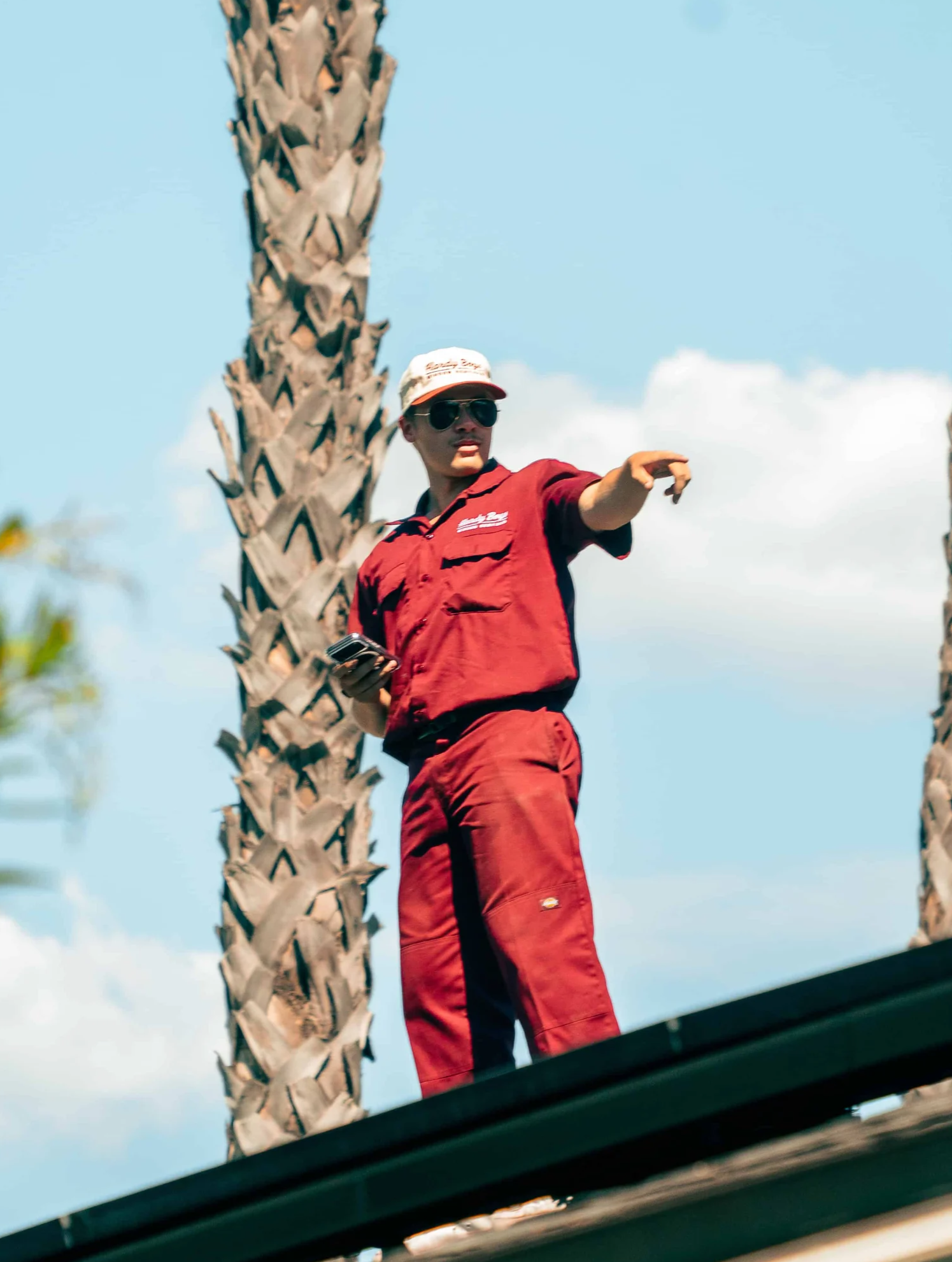 Man in red clothing and sunglasses points beside a palm tree under a blue sky.
