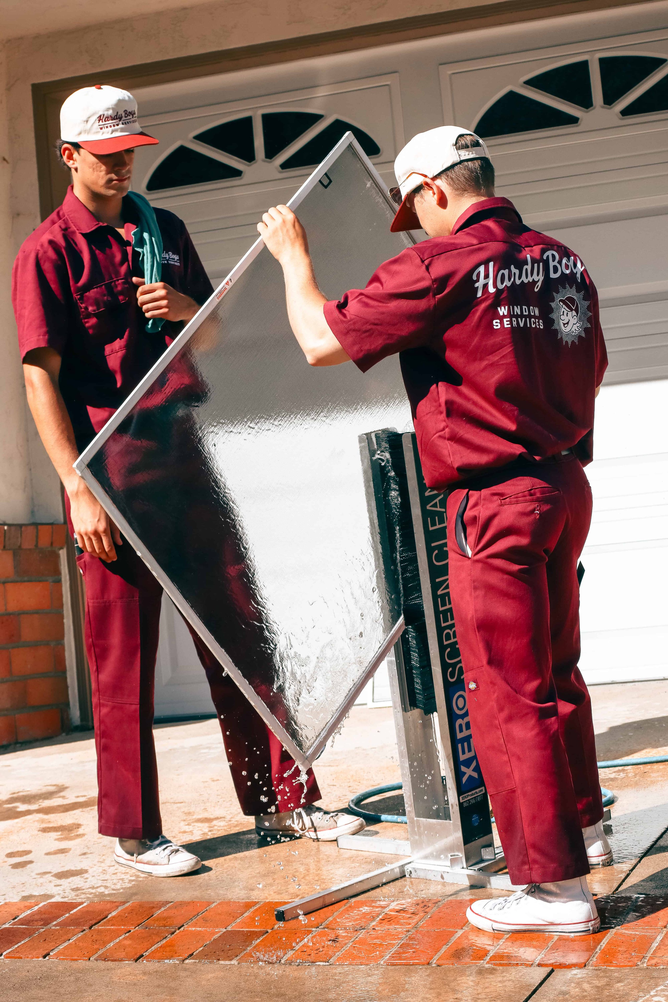 Two window-service workers clean a large wet window screen beside a garage door.