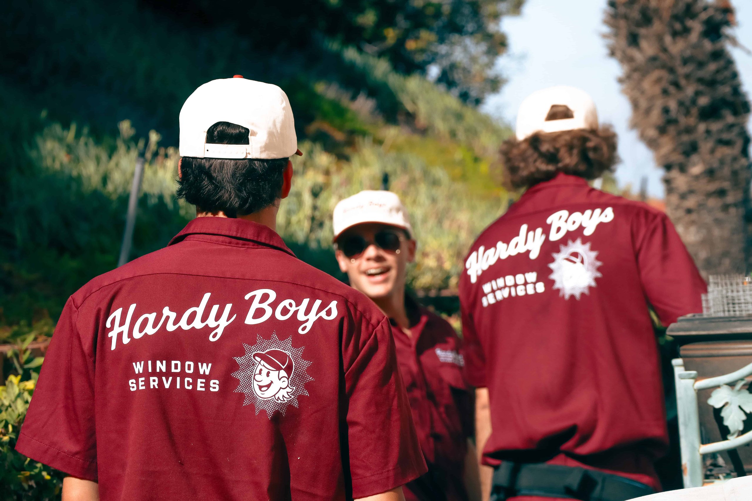 Bearded person in a white hard hat and burgundy shirt labeled “WINDOW SERVICES” outdoors.
