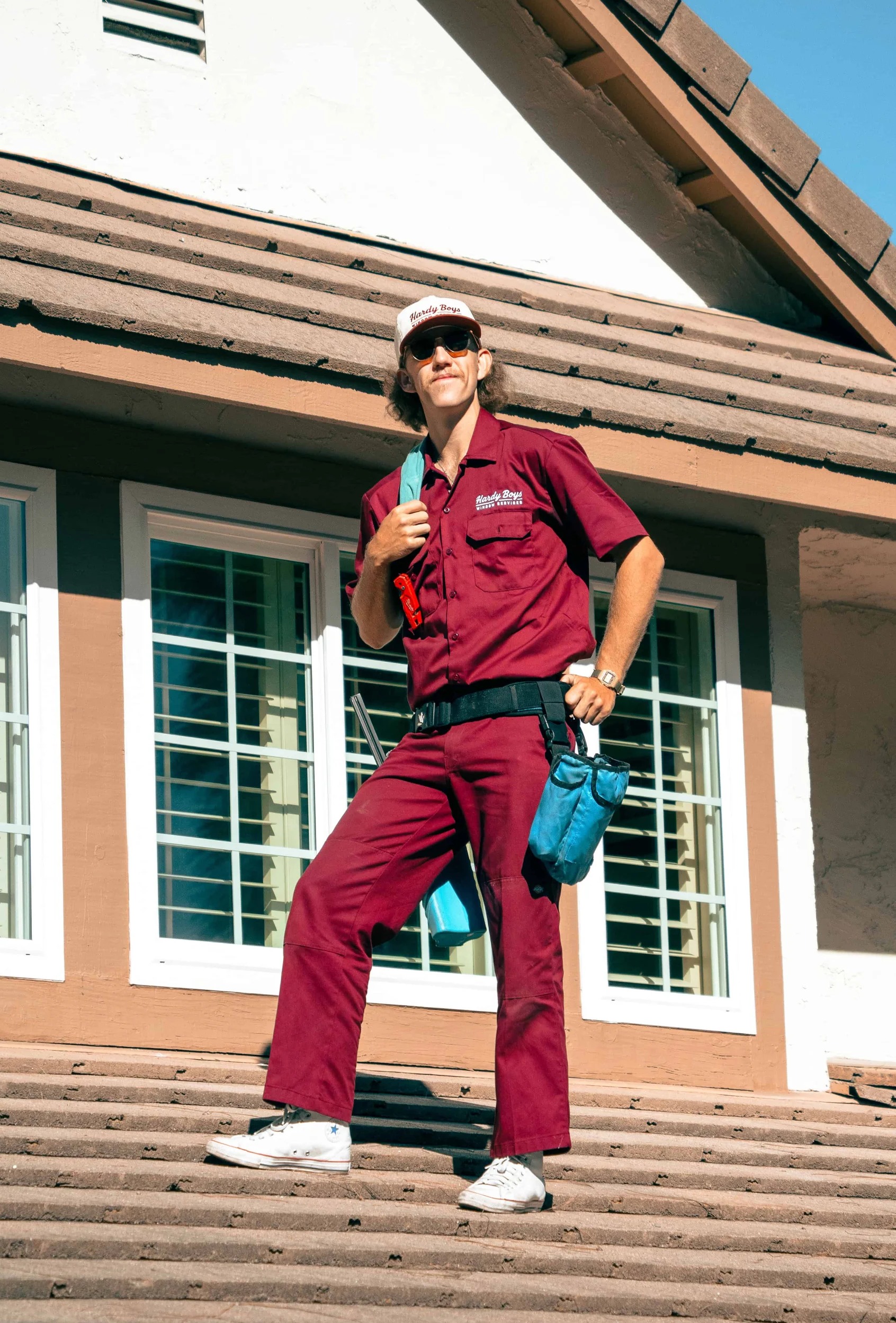 Worker in a burgundy uniform and sunglasses stands on a tiled roof with blue tool bags.